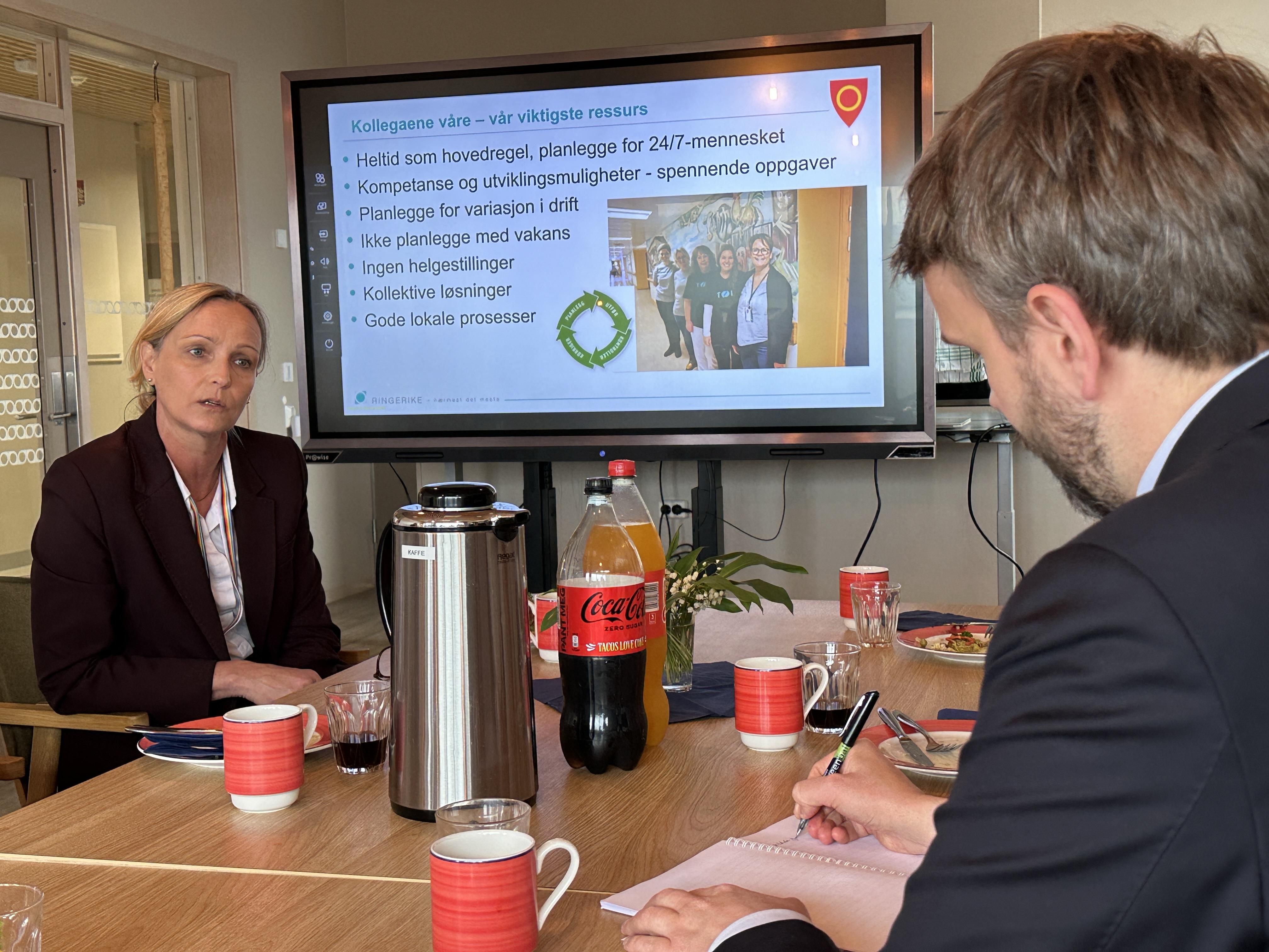 Kommunalsjef for Helse og omsorg snakker og viser en powerpoint presentasjon, mens statsr&aring;den lytter og noterer p&aring; en blokk.
